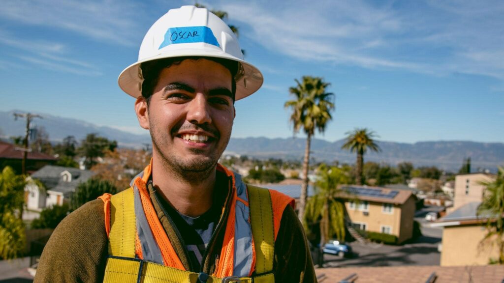 Oscar Flores on rooftop during solar installation job. View of residential community with palm trees behind him.